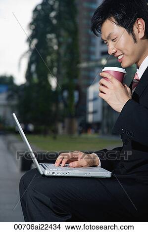 Picture - A man has a coffee as he sits and uses his laptop outdoors. Fotosearch