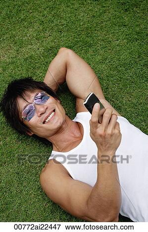 A man lies down on the grass and uses his cellphone View Large Photo Image Stock Photo - A man lies down on the grass and uses his cellphone. Fotosearch