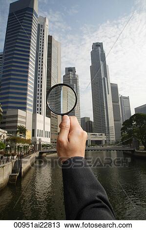Stock Image - Hand holding magnifying glass at skyscrapers. Fotosearch