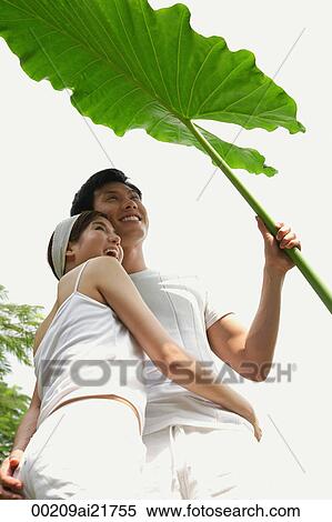 Stock Photography - Man and woman hugging and standing under large leaf. Fotosearch
