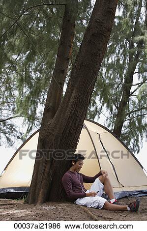 Stock Photograph - Man leaning against tree listening to music on MP3 player, tent in background, camping. Fotosearch