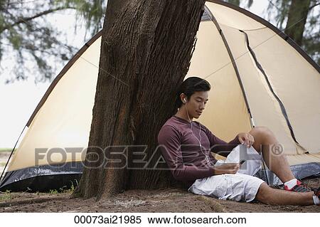 Stock Photography - Man leaning against tree listening to music on MP3 player, camping with tent. Fotosearch