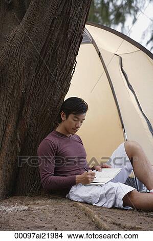 Picture - Man leaning against tree writing in journal, notebook, with tent in background, camping. Fotosearch