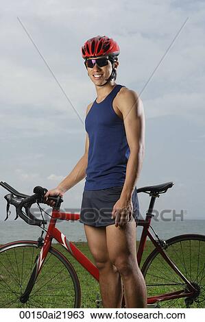 Stock Image - Man standing next to bike along ocean, looking at camera, smiling. Fotosearch