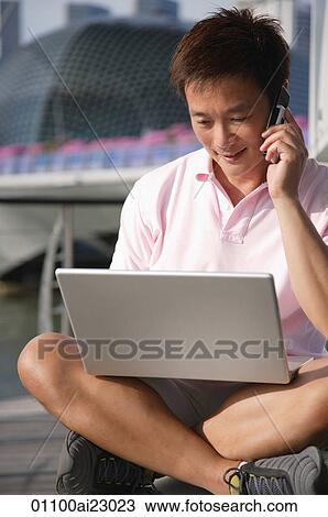 Stock Image - Man working on laptop while talking on the phone outdoors. Fotosearch