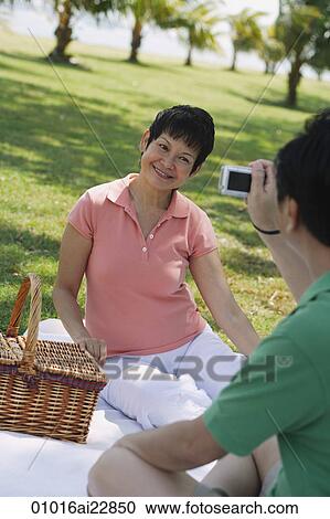 Stock Image - Mature couple having a picnic in the park and taking a picture. Fotosearch
