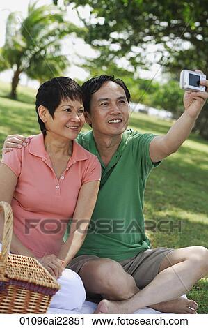 Stock Image - Mature couple having a picnic in the park and taking a picture of themselves. Fotosearch