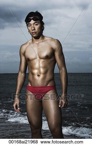 Stock Photo - Muscled man in swim trunks by ocean. Fotosearch