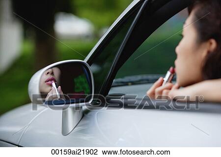 Stock Image - Woman applying lipstick in side view mirror of car, while in driver's seat. Fotosearch