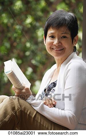 Stock Image - Woman relaxing with book in the garden and smiling at camera. Fotosearch