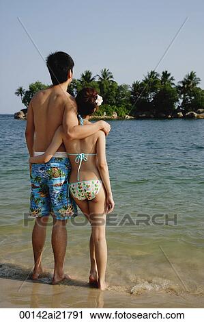 Stock Image - Young couple on beach, arms around each other, back to camera. Fotosearch