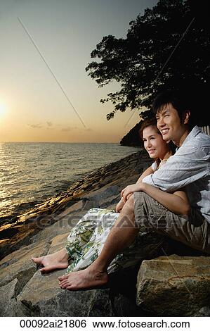 Stock Photograph - Young couple sitting on rocks at sunset looking out over the ocean. Fotosearch