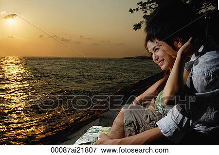 Stock Photo - Young couple sitting on rocks overlooking beach. Fotosearch