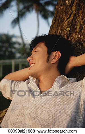 Stock Photo - Young man leaning against tree, relaxing. Fotosearch
