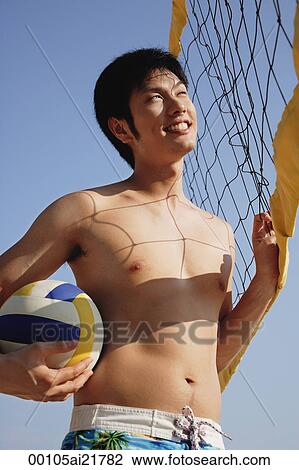 Stock Image - Young man on beach holding volleyball by net. Fotosearch