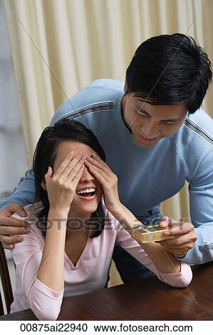 Stock Image - Young woman covering her eyes while man is giving her a present. Fotosearch