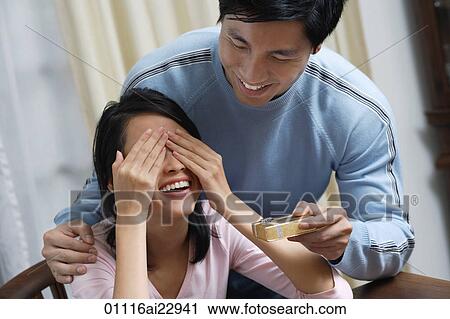 Stock Image - Young woman covering her eyes while man is giving her a present. Fotosearch