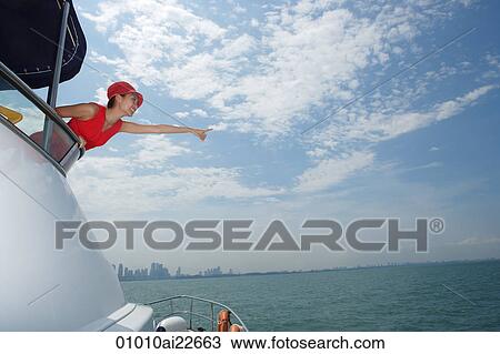 Stock Image - Young woman on yacht, pointing into distance. Fotosearch