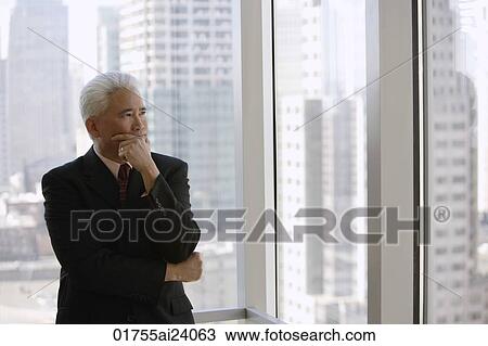 Stock Image - business man at window, hand on chin. Fotosearch