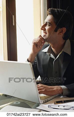 Stock Photo - business man working at laptop, hand on chin, looking out window. Fotosearch
