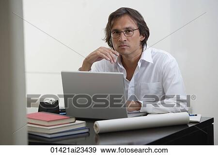 Stock Photo - Businessman using laptop at the desk, looking at camera. Fotosearch