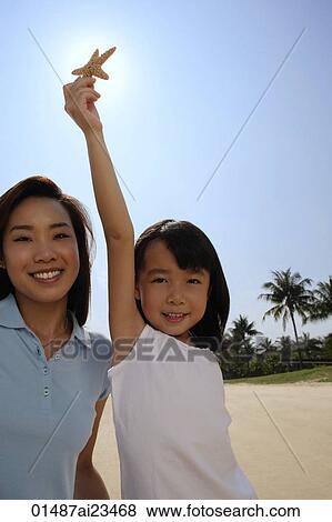 Stock Photo - Mother with daughter holding up shell at the beach, smiling at camera. Fotosearch