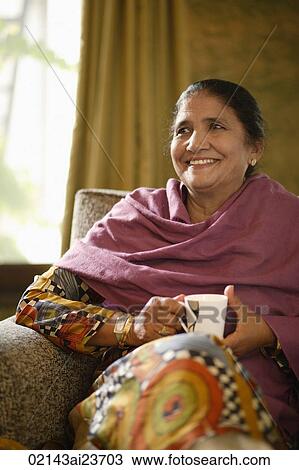 Stock Image - older woman sitting on couch, holding tea. Fotosearch