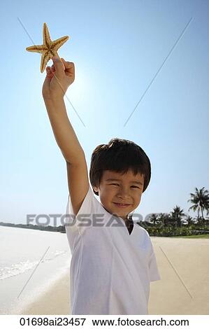 Young boy holding up shell at the beach, looking at camera View Large Photo Image Stock Photo - Young boy holding up shell at the beach, looking at camera. Fotosearch