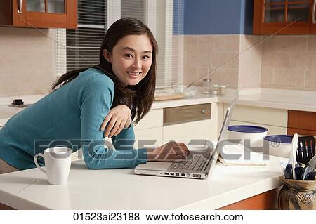 Stock Photo - Young woman using laptop on kitchen top, looking at camera. Fotosearch