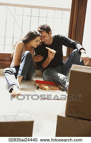Stock Image - young couple having pizza in new home, surrounded by boxes. Fotosearch