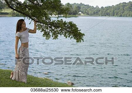 Stock Image - Woman standing under tree by lake. Fotosearch