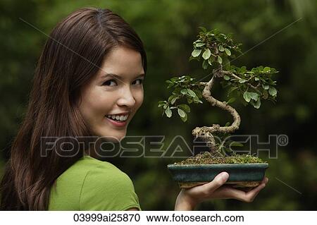 Stock Image - Young woman holding up bonsai tree. Fotosearch