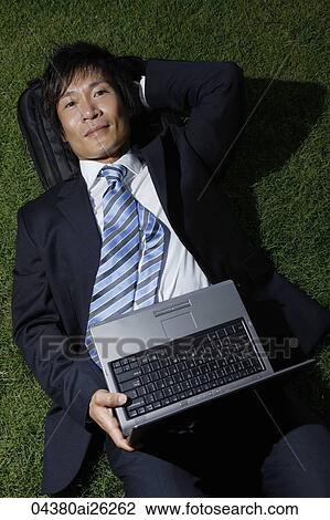 Stock Image - Businessman lying on grass with open laptop. Fotosearch