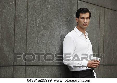 Stock Photo - "man in white shirt holding a cup of coffee, leaning against wall". Fotosearch