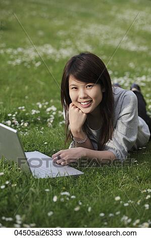 Stock Image - Chinese woman laying on grass with laptop.. Fotosearch