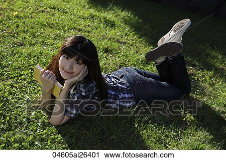 Stock Image - Asian woman laying on grass holding a book and smiling.. Fotosearch