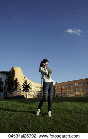 Stock Image - Asian woman talking on phone while standing on grass.. Fotosearch