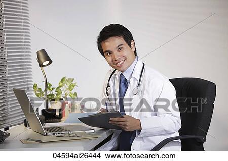 Doctor sitting at desk working on computer, smiling View Large Photo Image Picture - Doctor sitting at desk working on computer, smiling. Fotosearch