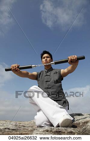 Stock Image - Chinese man holding nunchucks while sitting. Fotosearch
