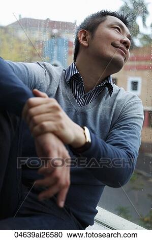 Stock Image - Chinese man looking out window and smiling at rain. Fotosearch