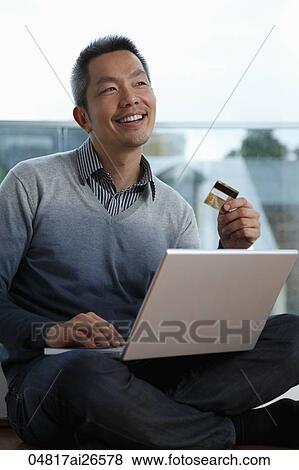 Stock Photo - Chinese man sitting near window with lap top and credit card. Fotosearch