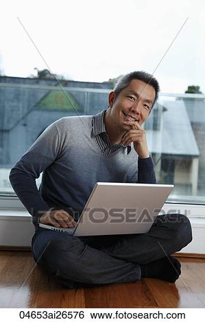 Stock Photograph - Chinese man sitting on floor by window with laptop. Fotosearch