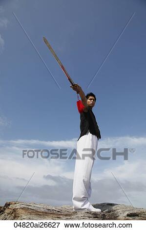 Stock Photo - Chinese man standing on a rock and pointing a sword up into the sky. Fotosearch