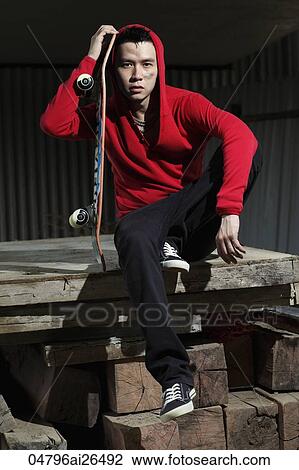 Stock Image - Chinese man wearing red hooded sweatshirt and holding skateboard.. Fotosearch