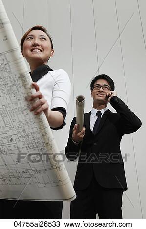 Stock Image - Chinese woman holding building plans while her male colleague talks on the phone.. Fotosearch