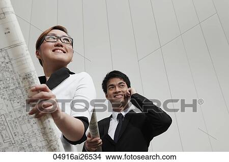 Picture - Chinese woman holding business plans while her male colleague talks on the phone.. Fotosearch