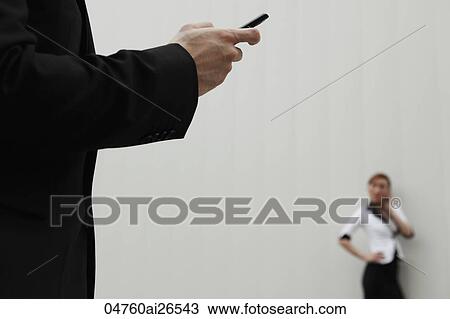 Stock Image - Close up of man using hand phone with woman talking on phone in background.. Fotosearch