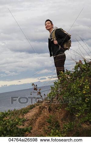 Stock Photo - Man wearing back pack and standing on trail over looking ocean. Fotosearch