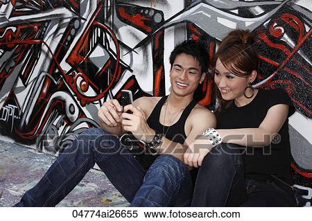 Young couple sitting on ground next to graffiti wall looking at hand phone View Large Photo Image Stock Photography - Young couple sitting on ground next to graffiti wall looking at hand phone. Fotosearch
