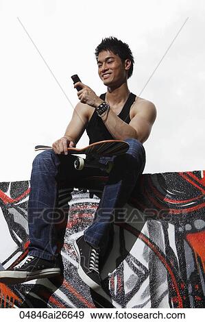 Young man sitting on wall with skateboard and holding phone View Large Photo Image Stock Photo - Young man sitting on wall with skateboard and holding phone. Fotosearch